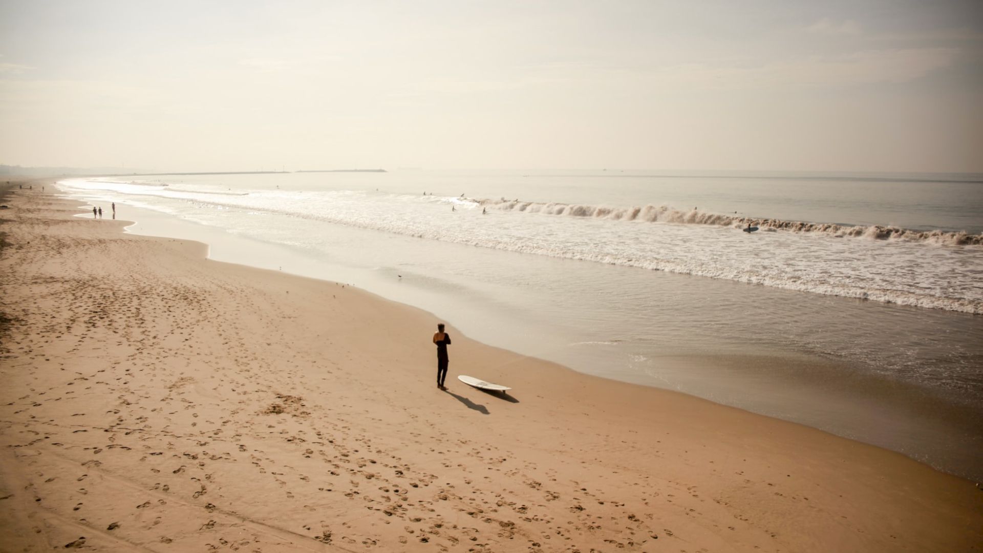A person stands on a sandy beach near the ocean, with a surfboard lying on the sand, under a clear sky.