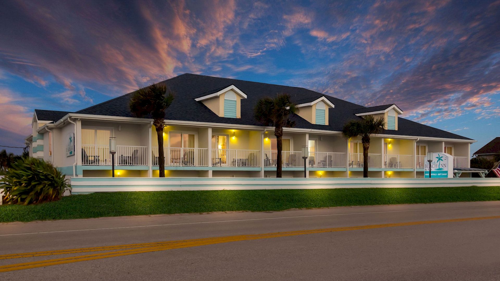A large house with a dark roof and white porch is lit up at dusk against a colorful sky, near a road.