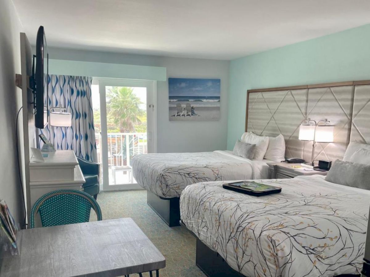 A hotel room with two beds, a wall-mounted TV, a desk, and a balcony view featuring palm trees. The decor is light and beach-themed.