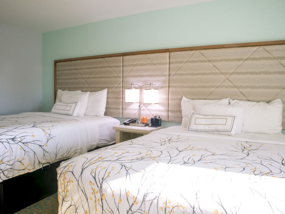 A hotel room features two double beds with white, patterned bedding, a central nightstand, and two lamps.