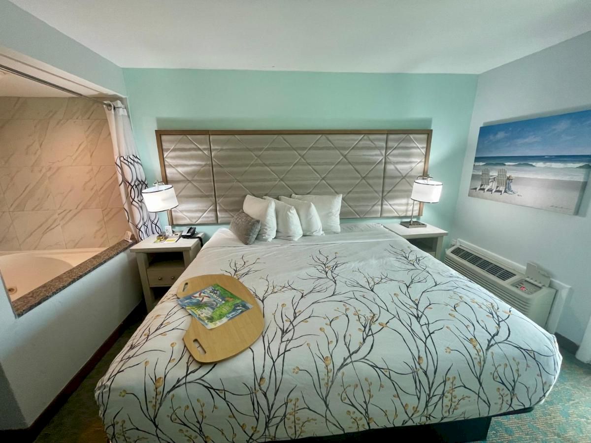 This image features a well-decorated bedroom with a king-sized bed, nightstands, lamps, a beach painting, and a visible bathroom area.