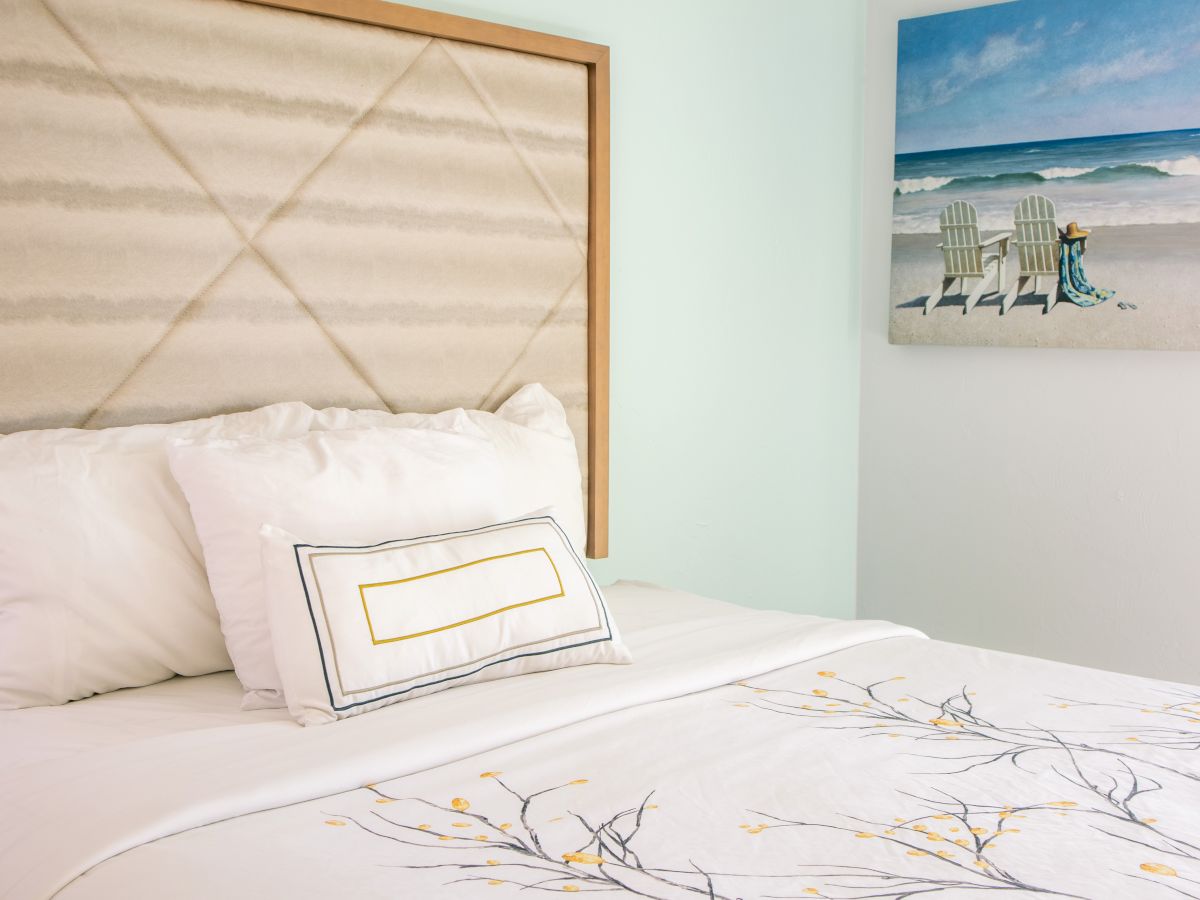A neatly made bed with a floral-patterned quilt, cushioned headboard, and a seaside painting on the wall.