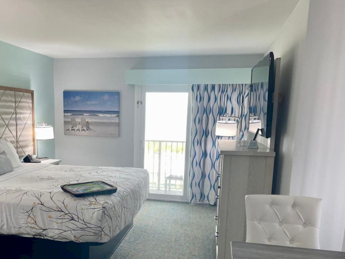 A tidy hotel room with a bed, wall art, TV, dresser, and balcony. The decor is modern and beach-themed, with shades of blue and white.