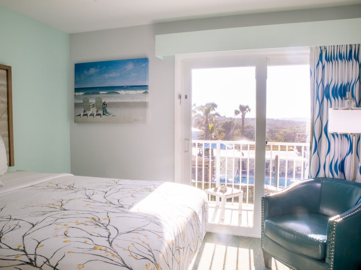 A bright hotel room with a bed, teal chair, beach painting, and a view of palm trees outside a window.