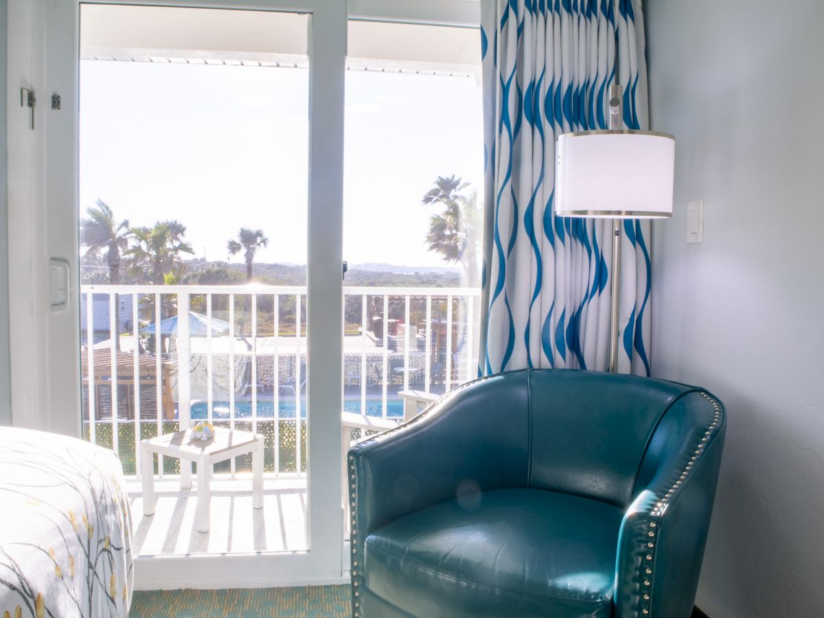 A room with a teal chair, lamp, and wavy curtains, featuring a balcony view with palm trees and a pool outside.