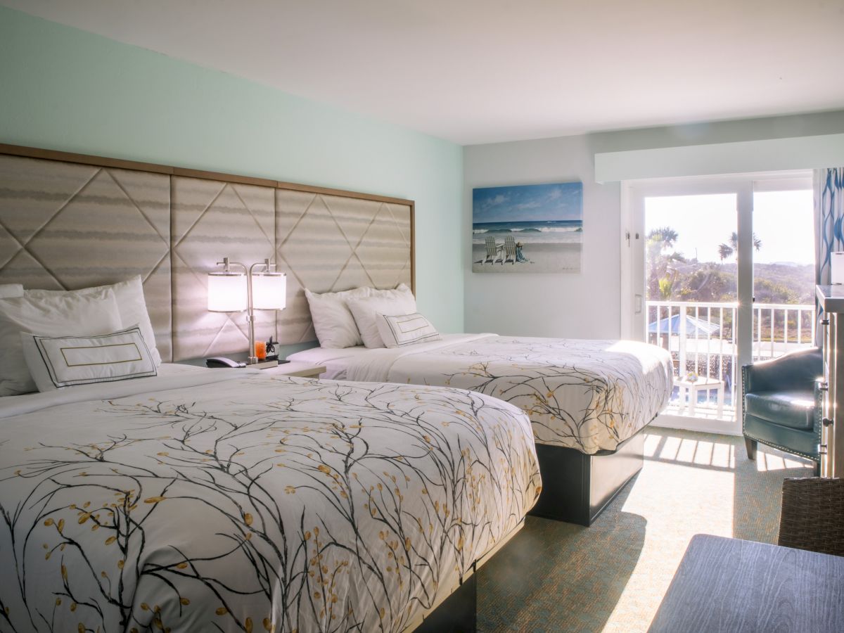 A bright hotel room with two beds, floral bedding, a lamp, a beach painting, and a balcony view, creating a relaxing atmosphere.