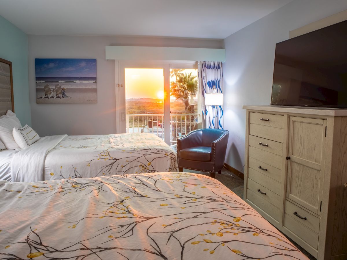 Hotel room with two beds, floral bedspread, wall art, balcony view of sunset, armchair, large TV, and dresser.