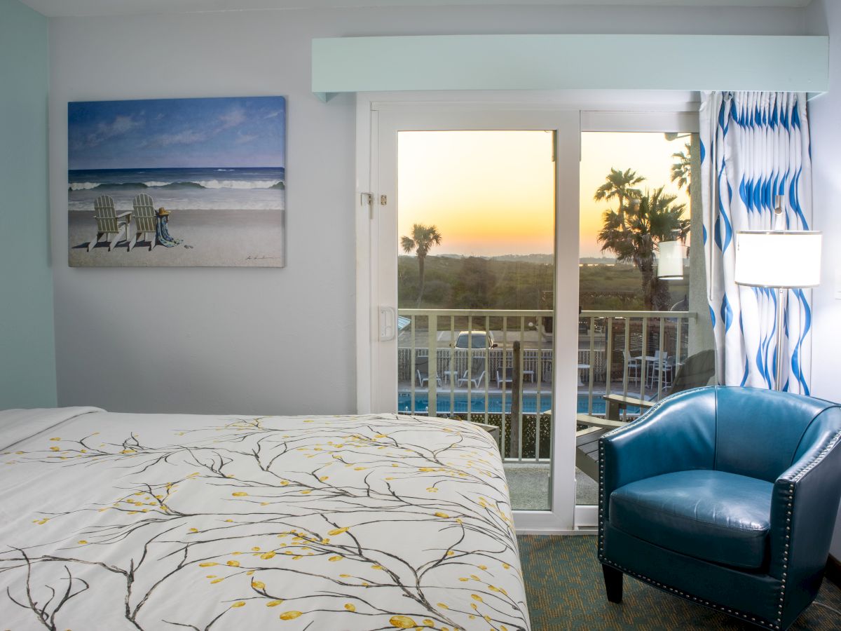 A cozy room with a patterned bedspread, blue chair, beach painting, and a sliding door opening to a sunset and a scenic balcony view.