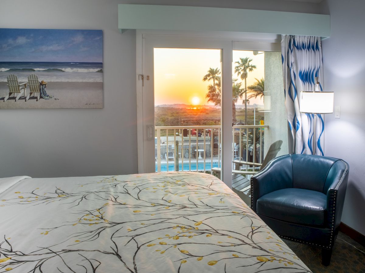 A cozy room with beach-themed decor, armchair, and a bed, overlooks a sunset view through sliding glass doors with trees visible outside.