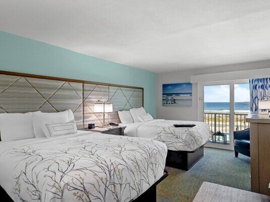Bright hotel room with two beds, floral bedding, ocean-view windows, a desk, and a balcony door.