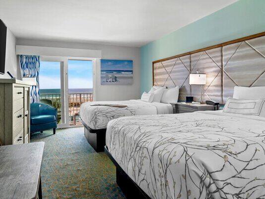 A bright hotel room with twin beds, ocean-view balcony doors, and teal walls.