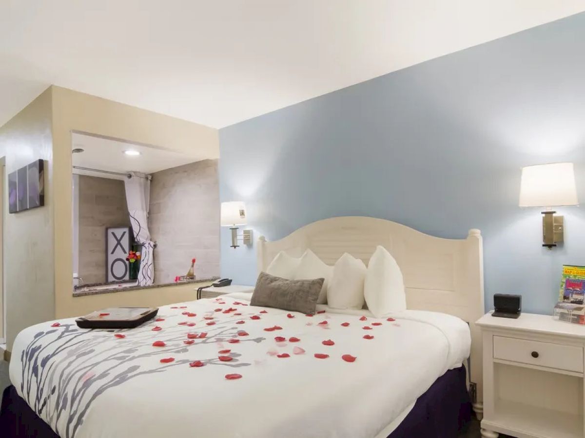 A cozy hotel room with a neatly made bed, adorned with rose petals, soft lighting, and a small sitting area in the background.