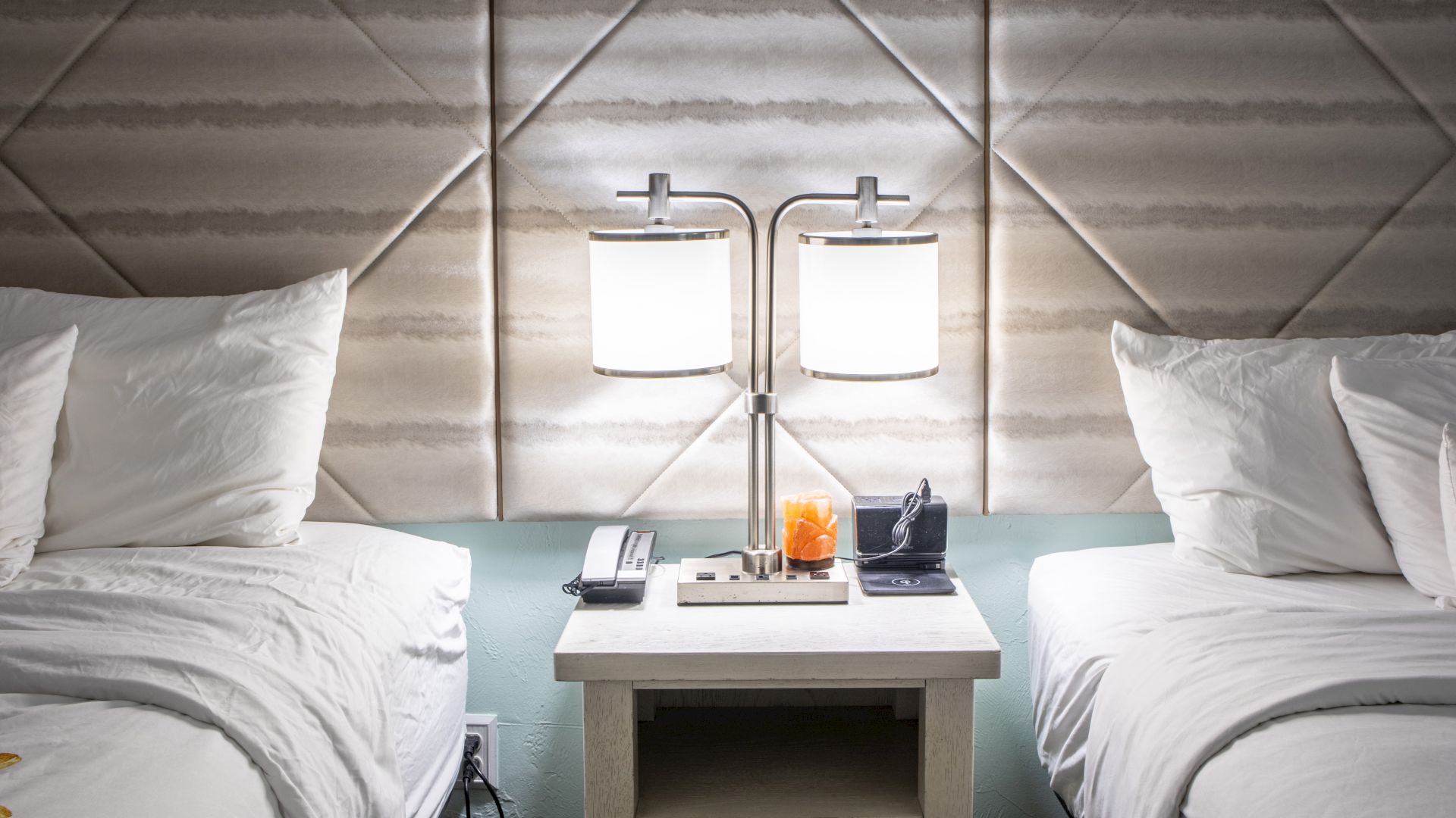 A hotel room with two beds, a dual lamp on a nightstand, and a phone, clock, and orange on the table in between.