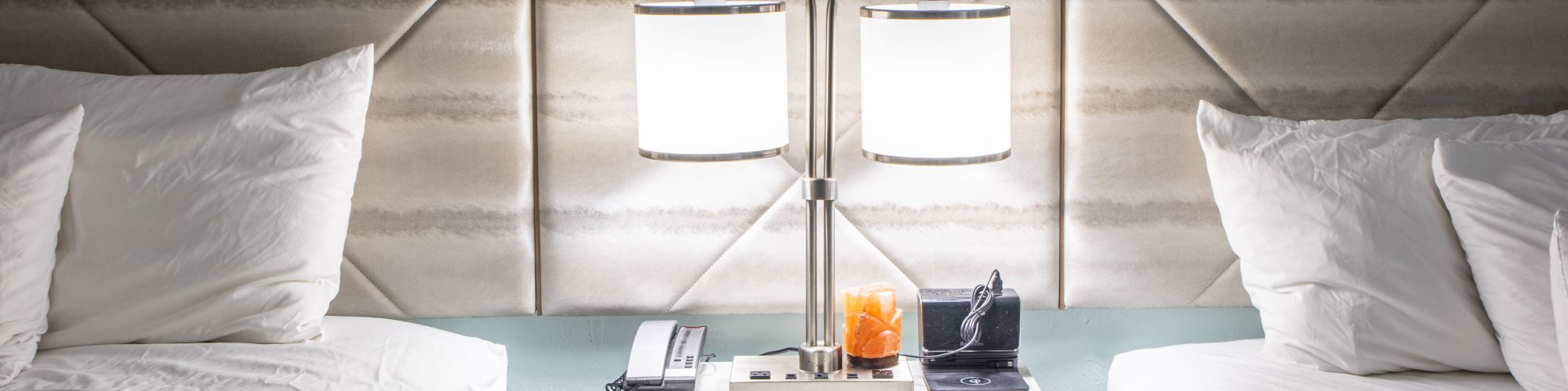 A hotel room with two beds, a dual lamp on a nightstand, and a phone, clock, and orange on the table in between.