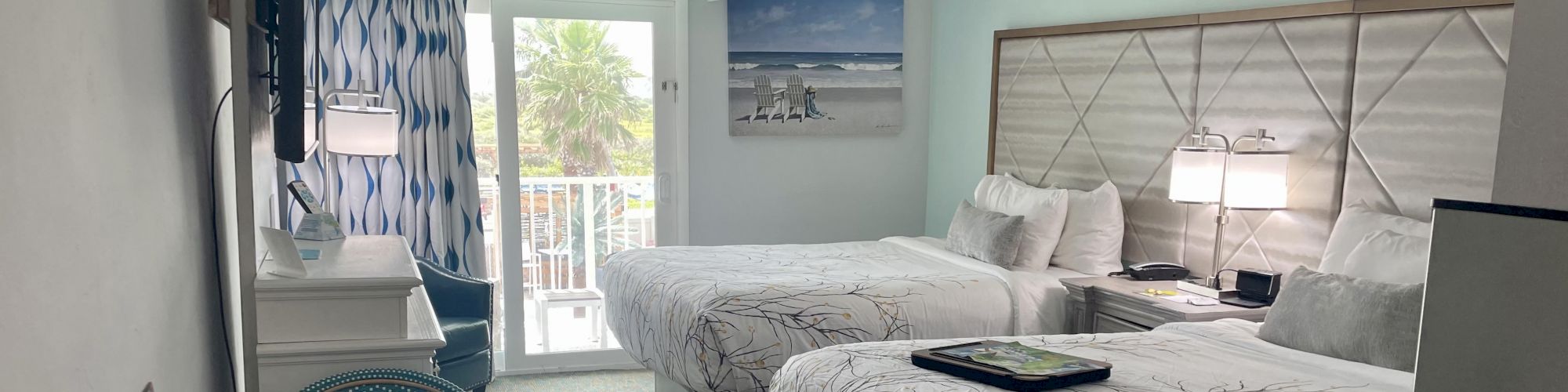 The image shows a hotel room with two beds, a wall-mounted TV, a desk, a picture of a beach on the wall, and a balcony with a view.