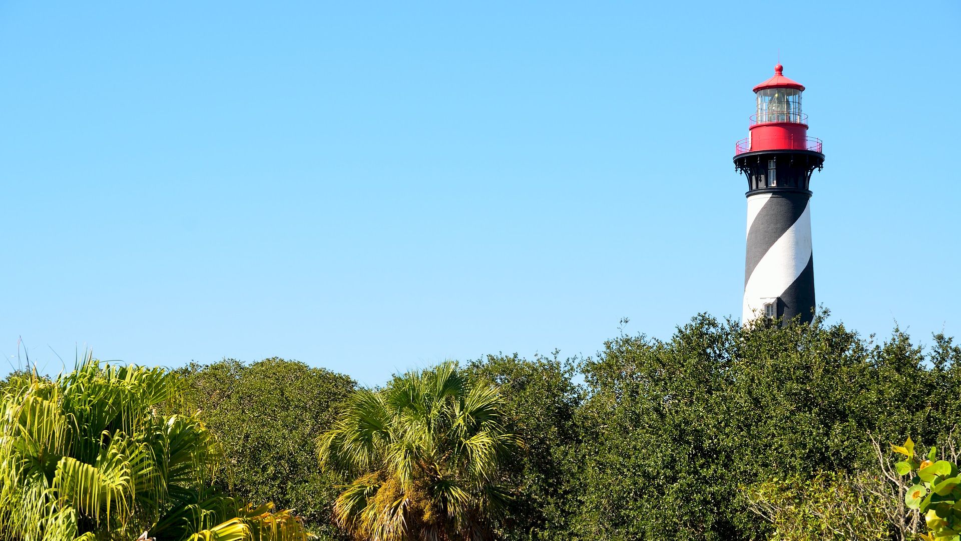 A black and white striped lighthouse with a red top stands behind lush greenery under a clear blue sky.