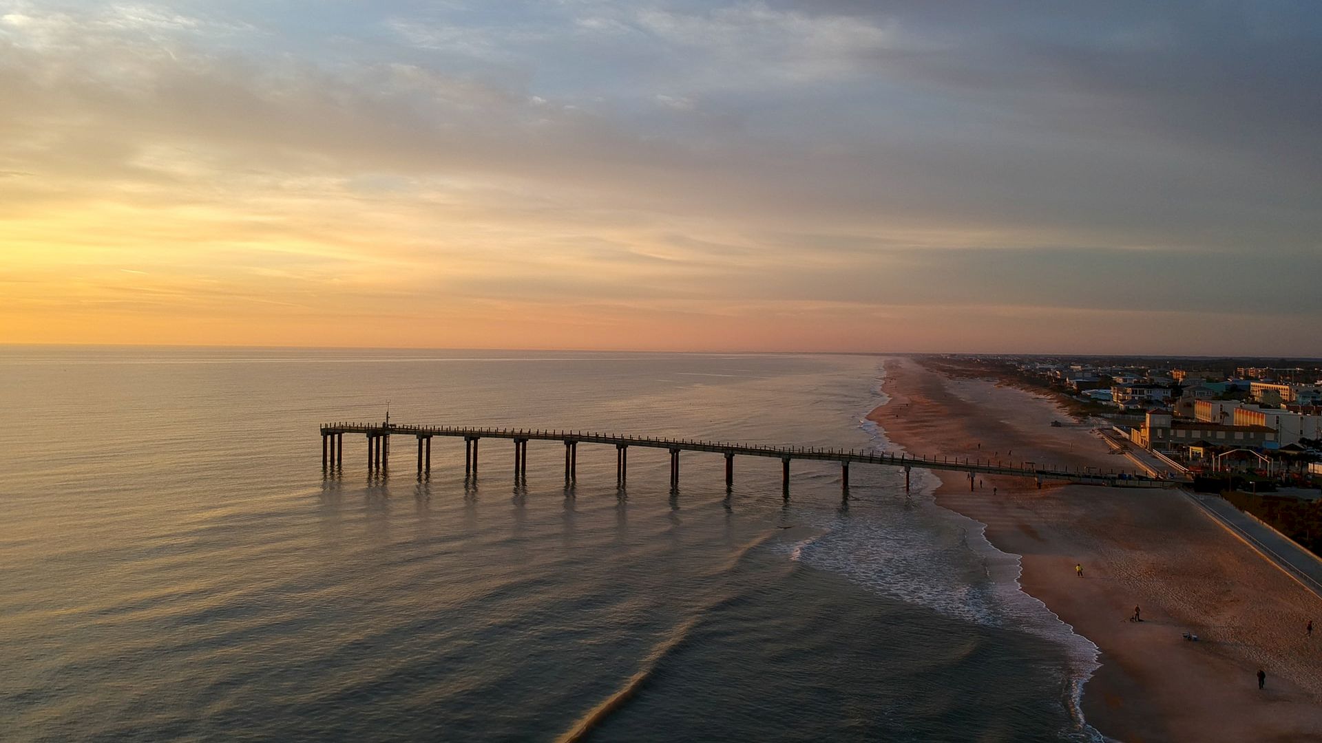 A scenic beach view at sunset with a long pier extending into calm ocean waters and buildings along the shoreline.