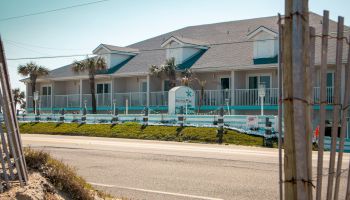 A two-story beachfront building with balconies, palm trees, and a road in the foreground on a sunny day.