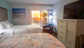 A cozy hotel room with two beds, patterned bedspreads, a large TV, and a balcony with a scenic sunset view, creating a relaxing ambiance.