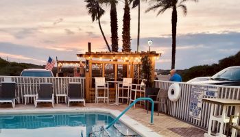 A poolside scene with lounge chairs, a decorated wooden bar, palm trees, and a sunset sky, creating a relaxing atmosphere.