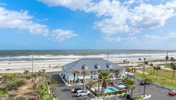 Beachfront building with parking lot, palm trees, and an outdoor pool, under a partly cloudy sky with sea waves in the background.