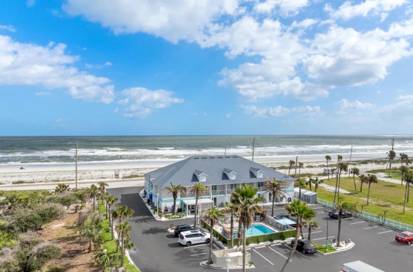 Beachfront building with parking lot, palm trees, and an outdoor pool, under a partly cloudy sky with sea waves in the background.