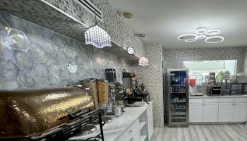 The image shows a modern kitchen or café area with hexagonal wall tiles, overhead lighting, a fridge, and coffee/tea machines on the counter.