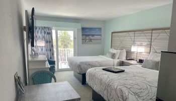 A hotel room with two beds, a television, a desk, and a balcony with a view. Decor includes beach-themed artwork.