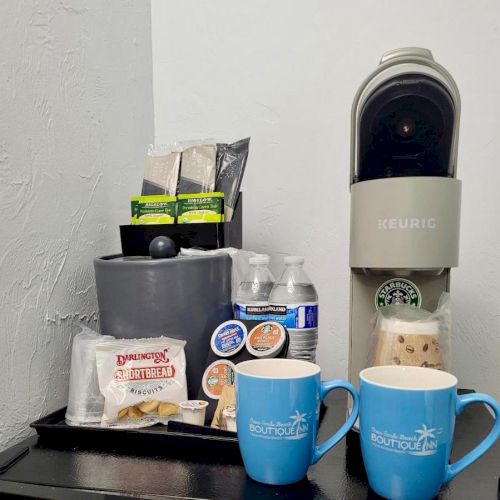 A coffee station with a Keurig machine, K-cups, mugs, tea bags, bottled water, and stir sticks.