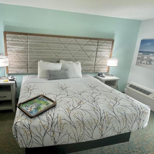 The image shows a neatly made bed in a hotel room with two nightstands, lamps, and a wall painting. A tray is placed on the bedspread.