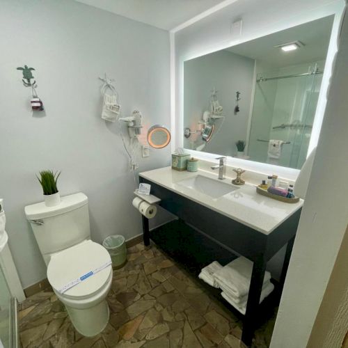 The image shows a modern bathroom with a toilet, sink, large mirror, and toiletries on a countertop. The floor has a tile design.