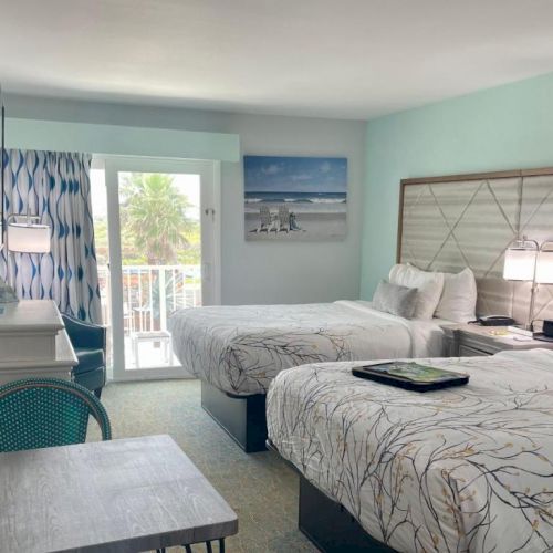 A hotel room with two beds, a TV, a desk, and a balcony with a view of palm trees. White and blue decor adds a fresh feel.