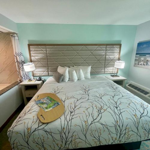 A cozy hotel room with a large bed, decorative pillows, lamps, and a scenic beach painting. There's an open view of a bathroom with a tub.