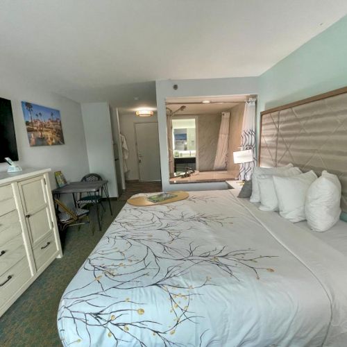 A cozy hotel room with a king bed, dresser, TV, and seating area, decorated with a nature-themed bedspread and soft lighting.