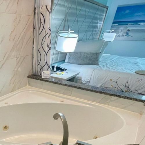 The image shows a luxurious bathroom with a white bathtub, marble tiles, and a view into a bedroom with a bed and beach-themed artwork.