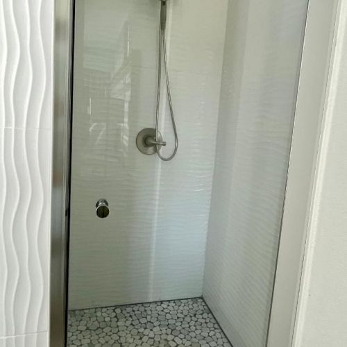 A modern shower with a glass door, white textured wall tiles, and a pebble stone floor.