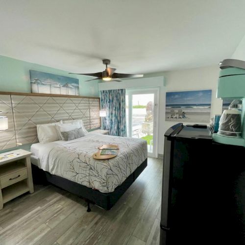 A cozy hotel room with a king bed, nightstands, modern decor, and a kitchenette, featuring soft teal walls and a view through glass doors.