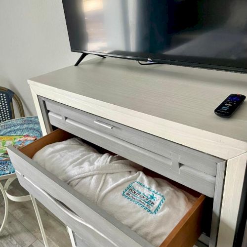 A TV sits on a dresser with an open drawer containing a folded bathrobe and a packet of wipes. A remote is on top of the dresser.