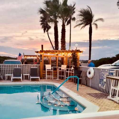 A poolside setting with lounge chairs, a tiki bar, string lights, and palm trees at sunset, creating a relaxing atmosphere.