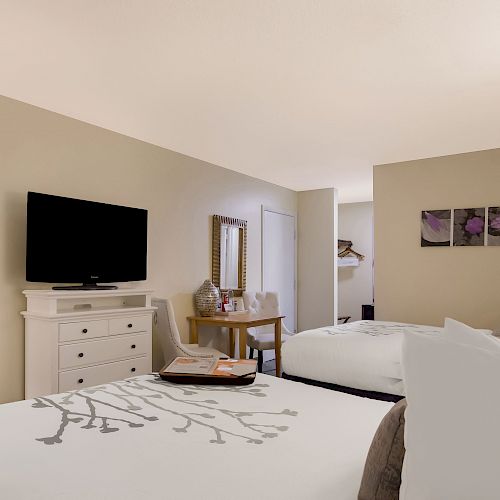 The image shows a hotel room with two beds, a TV, a dresser, a mirror, a lamp, and a small table with chairs and wall art.