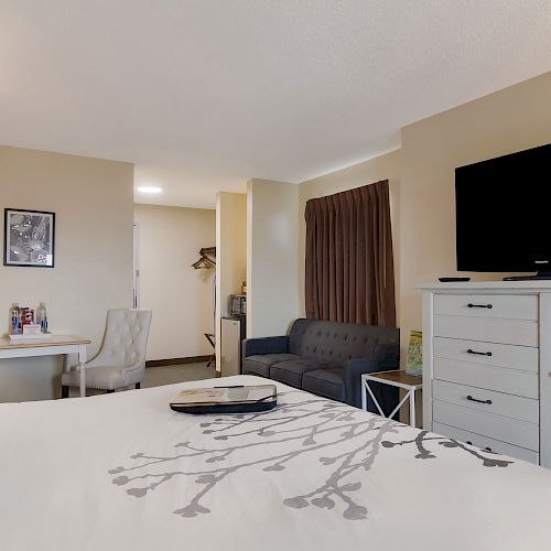 A hotel room with a bed, TV, dresser, small dining area, and a sofa. The decor is modern with neutral tones.