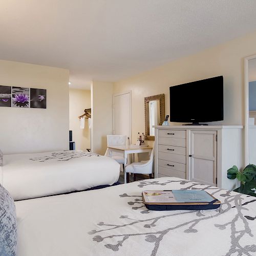 The image shows a hotel room with two beds, a flat-screen TV, dresser, vanity desk with chair, mirror, and decorative wall art above the beds.