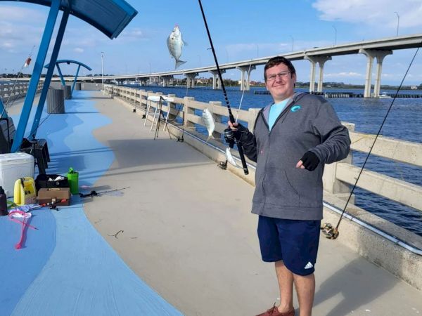 A person on a pier holds a fishing rod with a fish near a bridge and water on a sunny day, with fishing gear nearby.