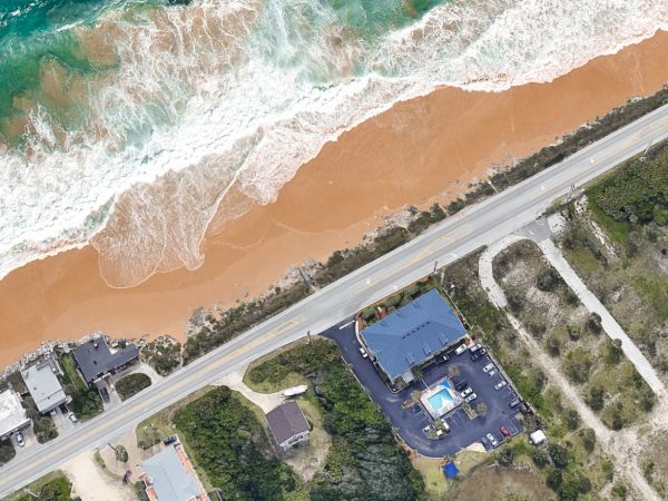 This image shows an aerial view of a coastline, with waves, a road, buildings, and a parking lot adjacent to the beach.