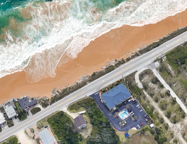 Aerial view of a coastline with waves, a road parallel to the shore, and a few buildings, including one with a pool.