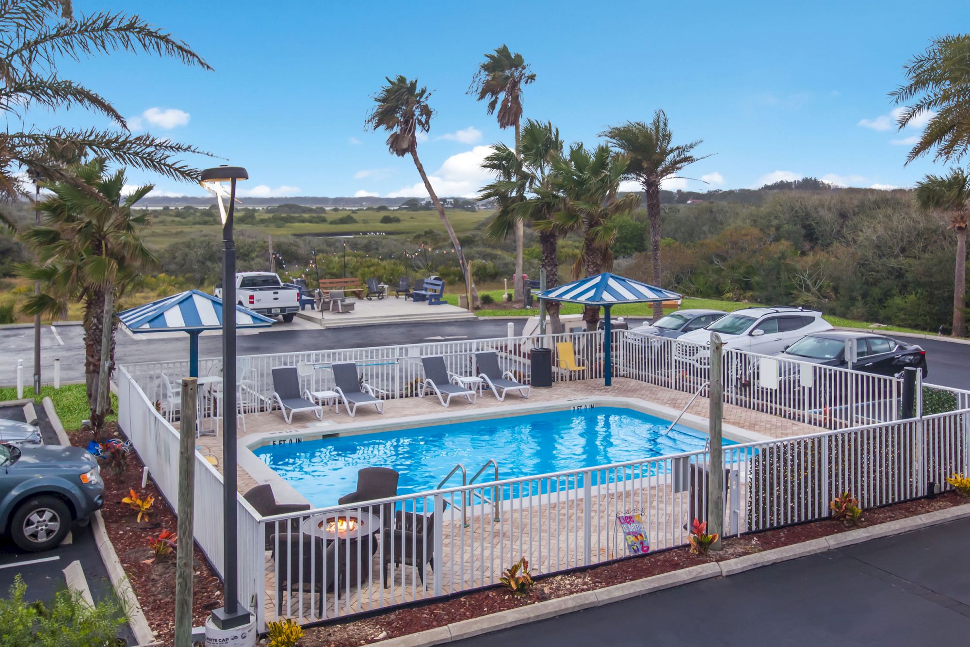 An outdoor swimming pool surrounded by a white fence, sun loungers, parked cars, palm trees, and blue sky with scattered clouds in the background.