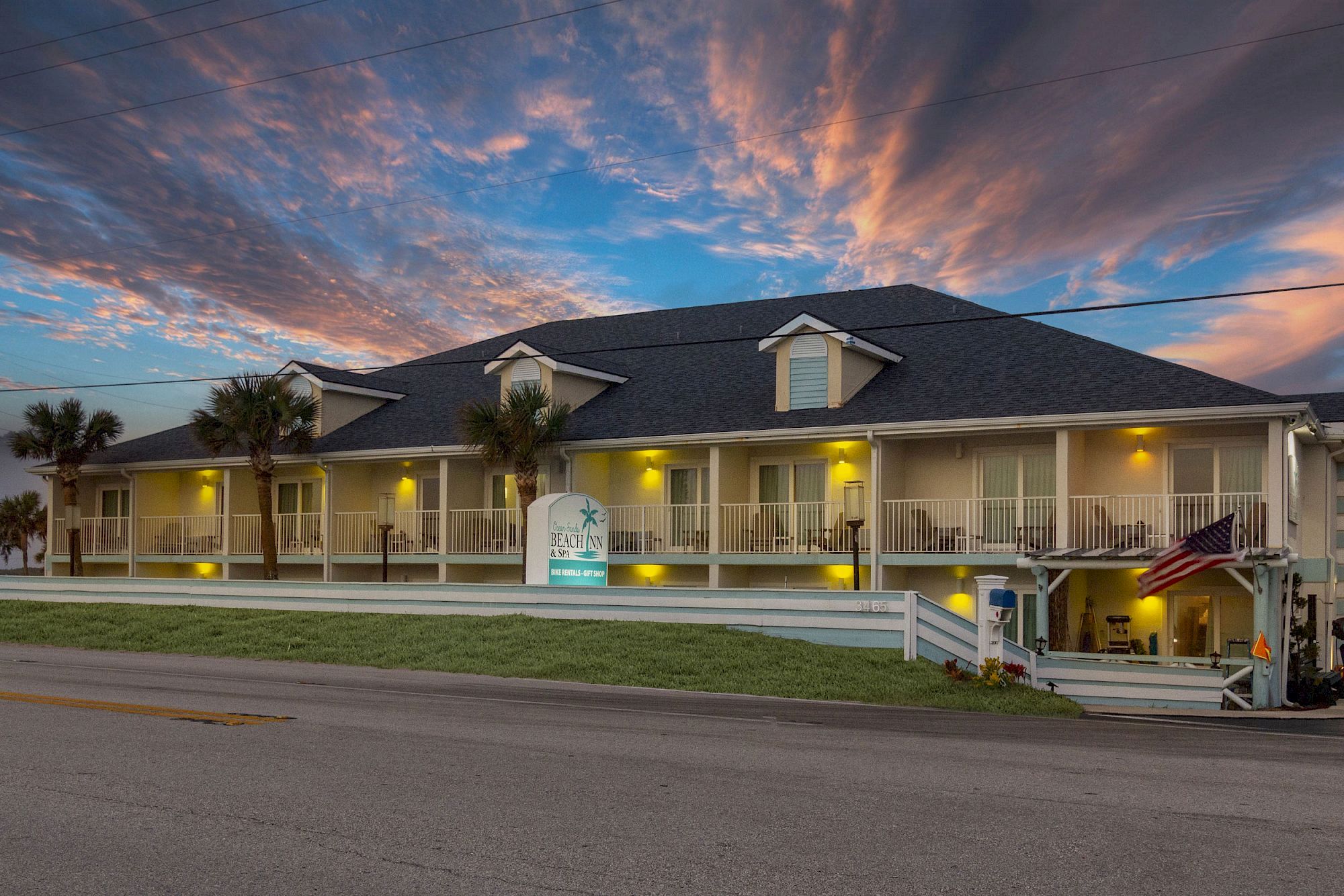 A two-story building with a "Beach House" sign, surrounded by a grassy area, illuminated by outdoor lights under a dramatic sunset sky.