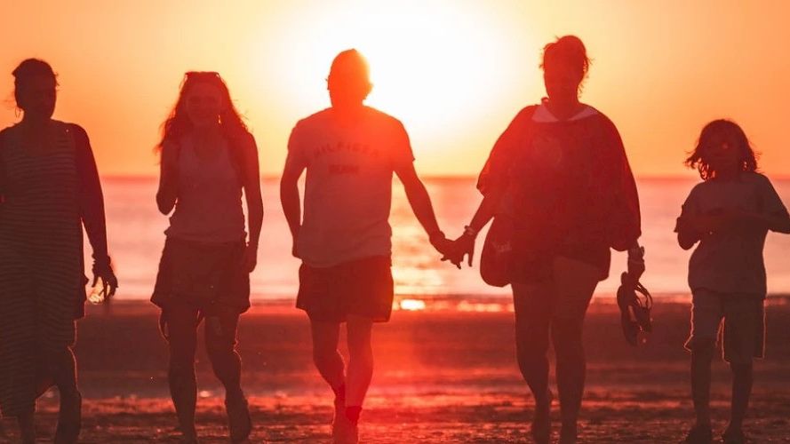 A group of people walking on a beach during sunset, silhouetted against the orange sky.
