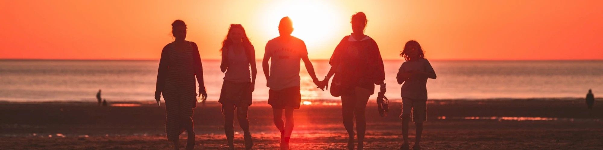 A group of people walking on a beach during sunset, silhouetted against the orange sky.