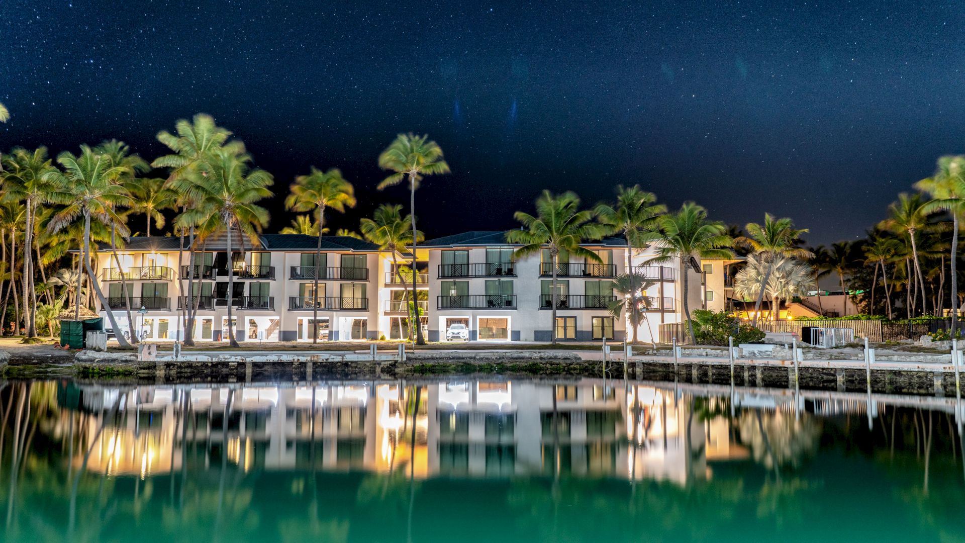 A luxurious, well-lit waterfront hotel is surrounded by palm trees, reflected in calm water under a starry night sky.